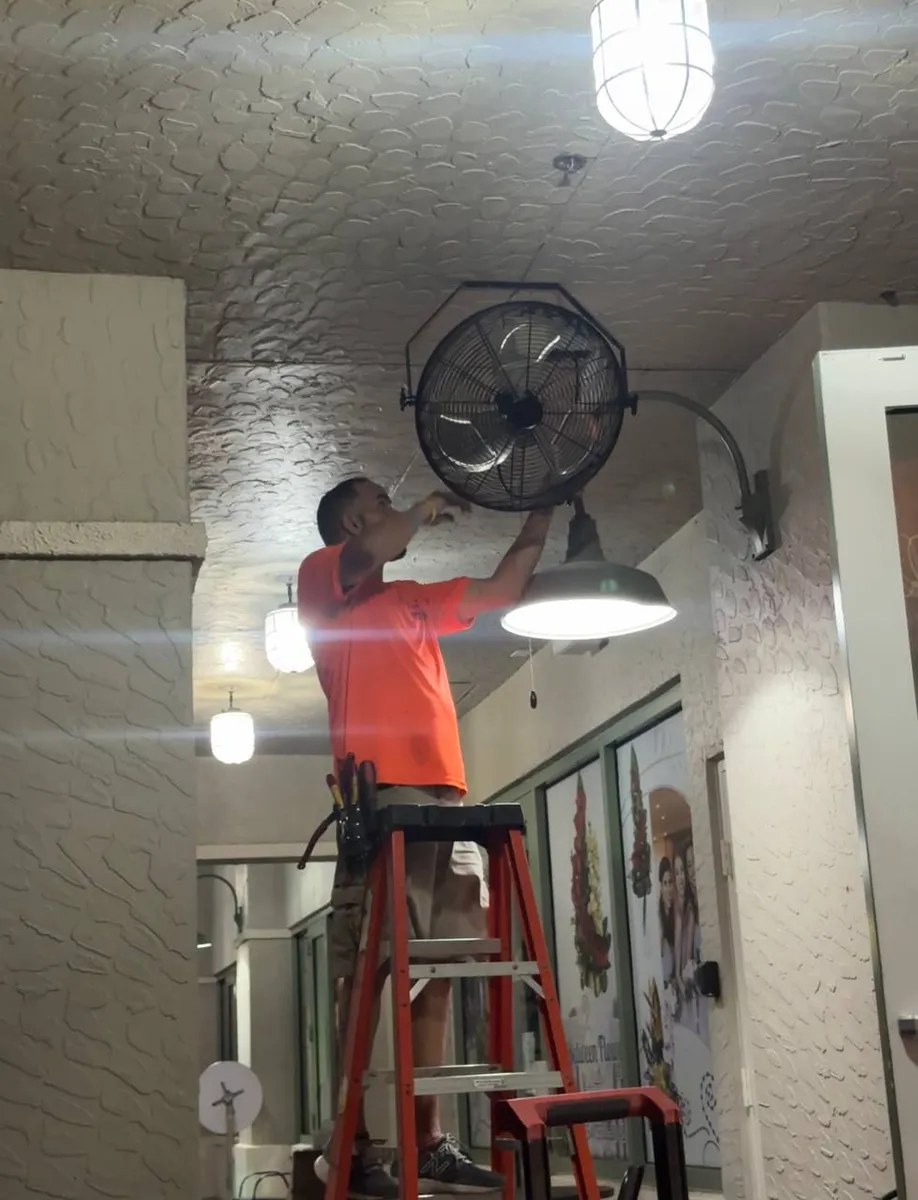Electrician on a ladder installing a fan next to a globe pendant light